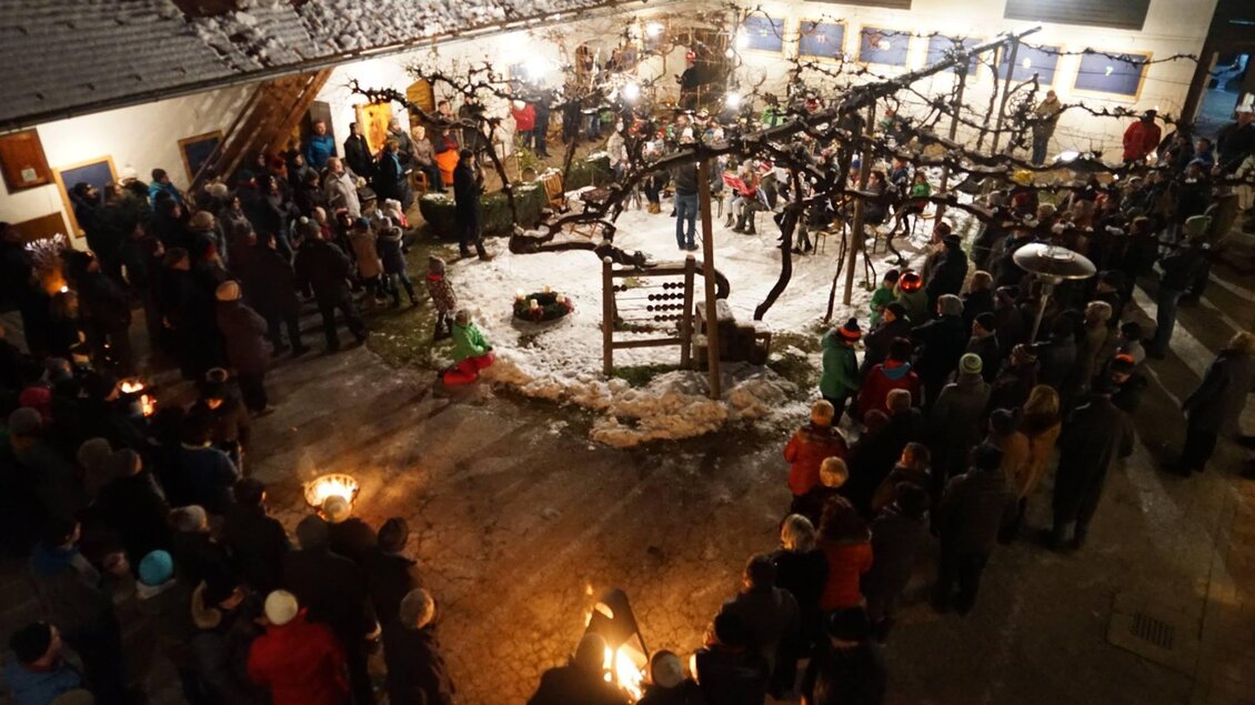 Eine festliche Versammlung in einem beleuchteten Innenhof, umgeben von schneebedeckten Bäumen. Menschen stehen zusammen und halten Fackeln, während sie die festliche Stimmung genießen.