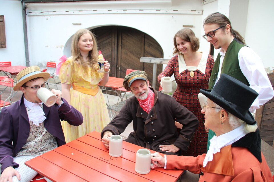 Darsteller in historischen Kostümen sitzen und stehen mit Krügen an einem Tisch bei den Schloss-Spielen Rheintal. | © Schloss Spiele Rheinthal