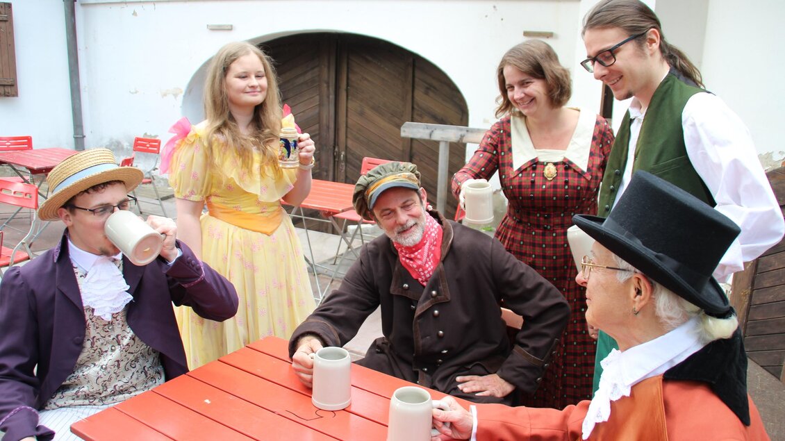 Darsteller in historischen Kostümen sitzen und stehen mit Krügen an einem Tisch bei den Schloss-Spielen Rheintal. | © Schloss Spiele Rheinthal