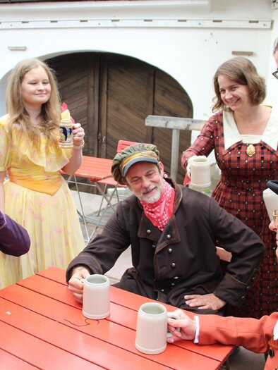 Darsteller in historischen Kostümen sitzen und stehen mit Krügen an einem Tisch bei den Schloss-Spielen Rheintal. | © Schloss Spiele Rheinthal