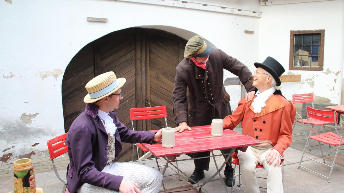 Drei Darsteller in historischen Kostümen spielen eine Szene an einem Tisch bei den Schloss-Spielen Rheintal. | © Schloss Spiele Rheinthal