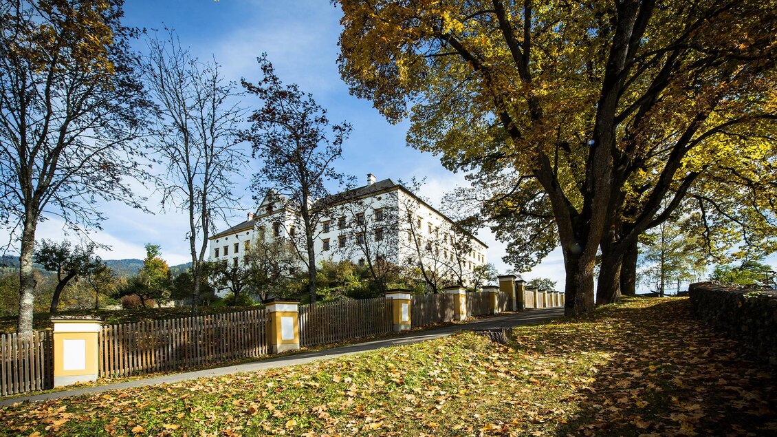 Ein historisches Gebäude umgeben von bunten Herbstbäumen. Der Weg ist mit fallenden Blättern gesäumt und der Himmel ist strahlend blau.