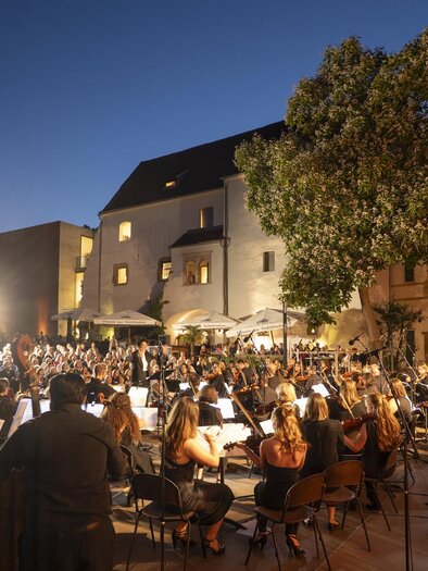 Ein großes Orchester spielt im Freien vor einem historischen Gebäude. Die Zuschauer sitzen in einem aufregenden Ambiente bei Dämmerlicht. | © Bernhard Bergmann