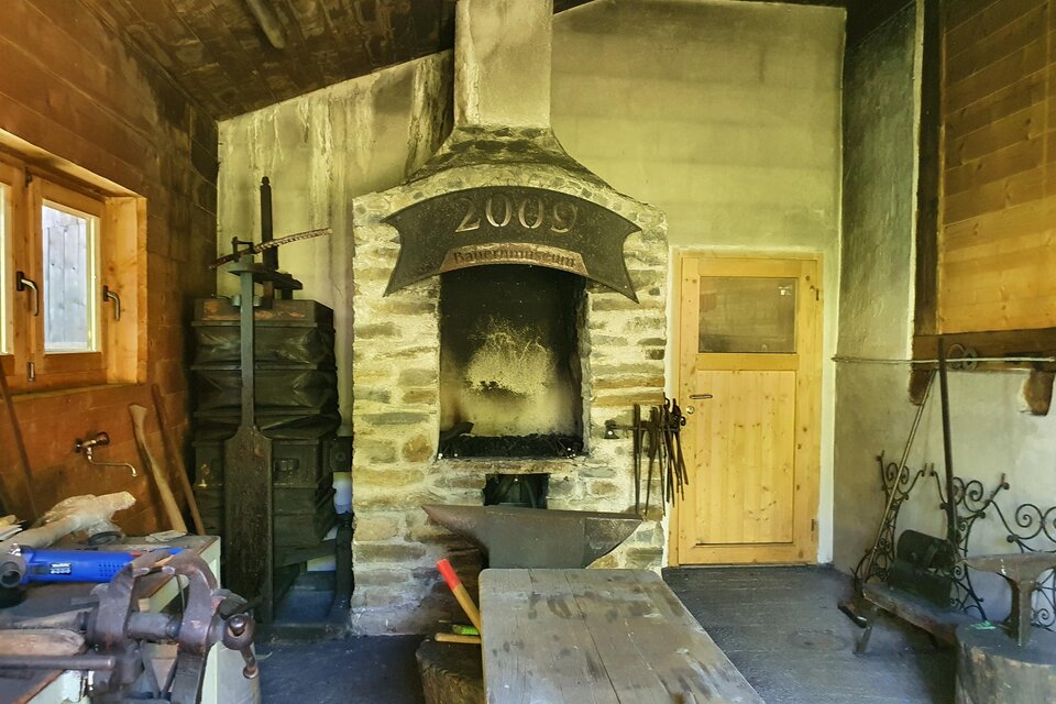 Eine alte Schmiede mit einem großen Ofen und verschiedenen Werkzeugen. Die Wände sind aus Holz und Stein, und die Atmosphäre ist rustikal.