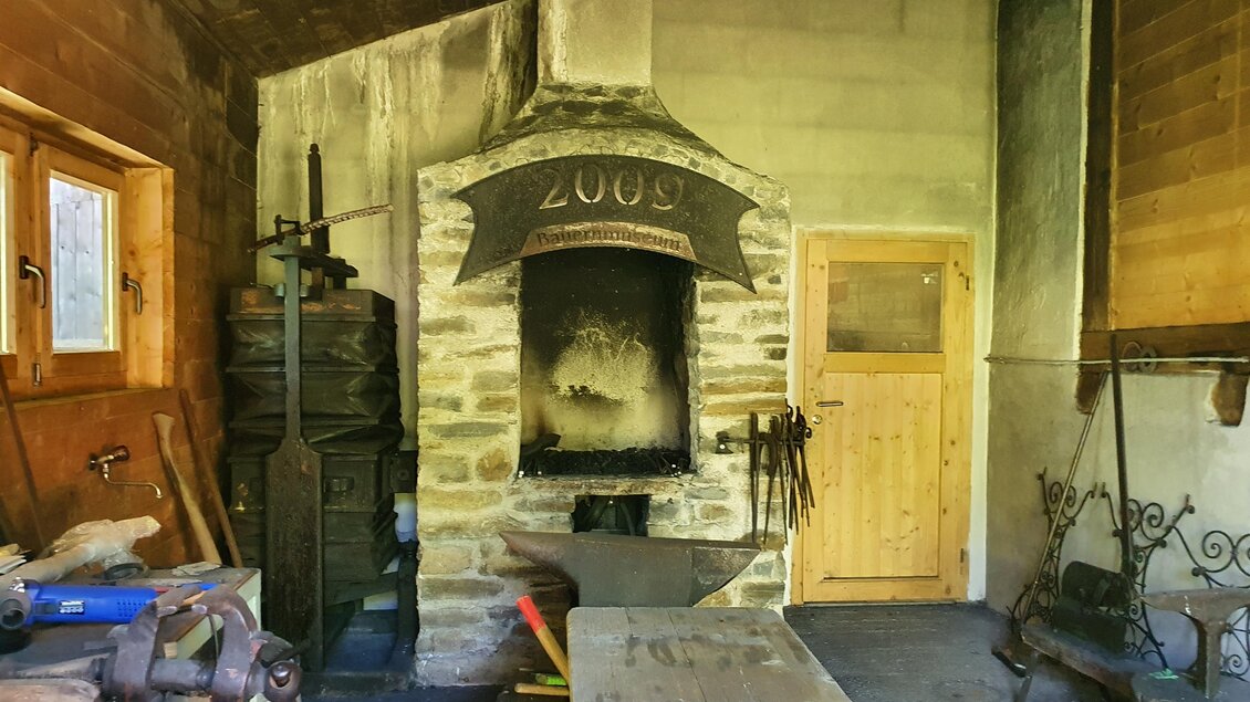 Eine alte Schmiede mit einem großen Ofen und verschiedenen Werkzeugen. Die Wände sind aus Holz und Stein, und die Atmosphäre ist rustikal.