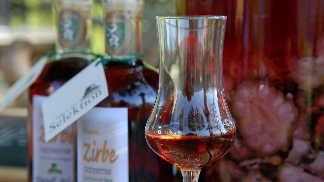 Ein Schnapsglas mit rot-braunem Zirbenschnaps, dahinter zwei Flaschen mit selbstgebranntem Schnaps und daneben ein Gefäß mit angesetztem Zirbenschnaps. | © Alfred Pohn