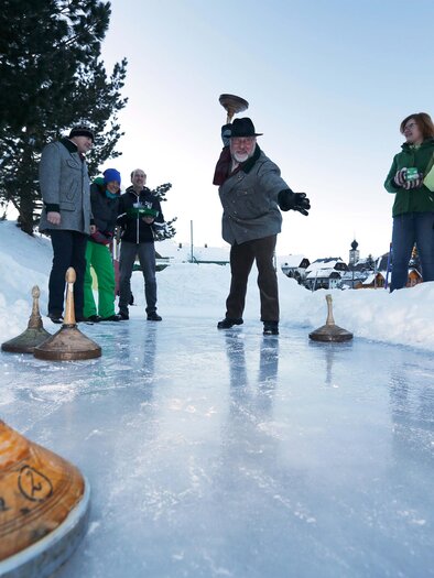 Eine Gruppe von Menschen spielt auf einem gefrorenen Teich im Schnee. Es sind verschiedene Holzgeräte zu sehen, die für das Spiel verwendet werden. | © Herbert Raffalt