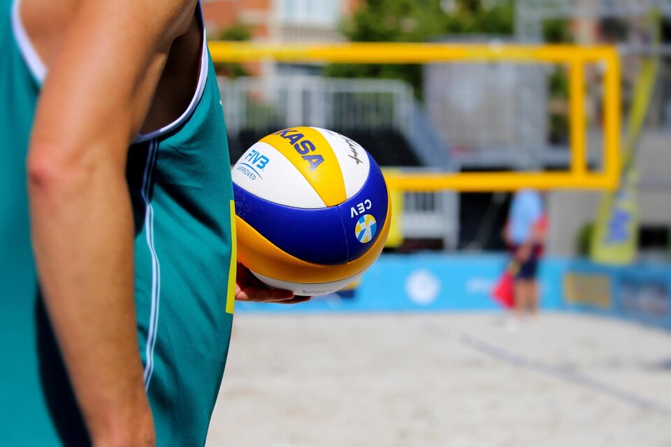 Ein Sportler hält einen Beachvolleyball in der Hand. Im Hintergrund ist ein Beachvolleyballfeld zu sehen. | © Pixabay