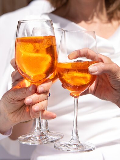 Three glasses with orange drinks are being clinked. In the background, people in light dresses can be seen. | © Pixabay