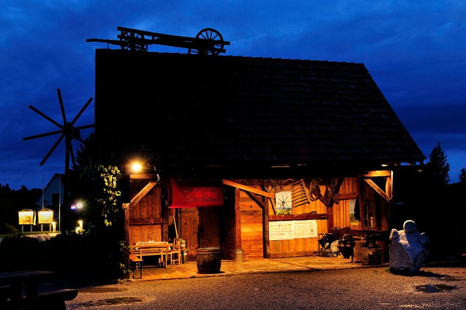 Ein rustikales Holzgebäude bei Nacht, beleuchtet von warmem Licht. Im Hintergrund ist ein Windrad und der Himmel ist dunkelblau. | © SPhotography