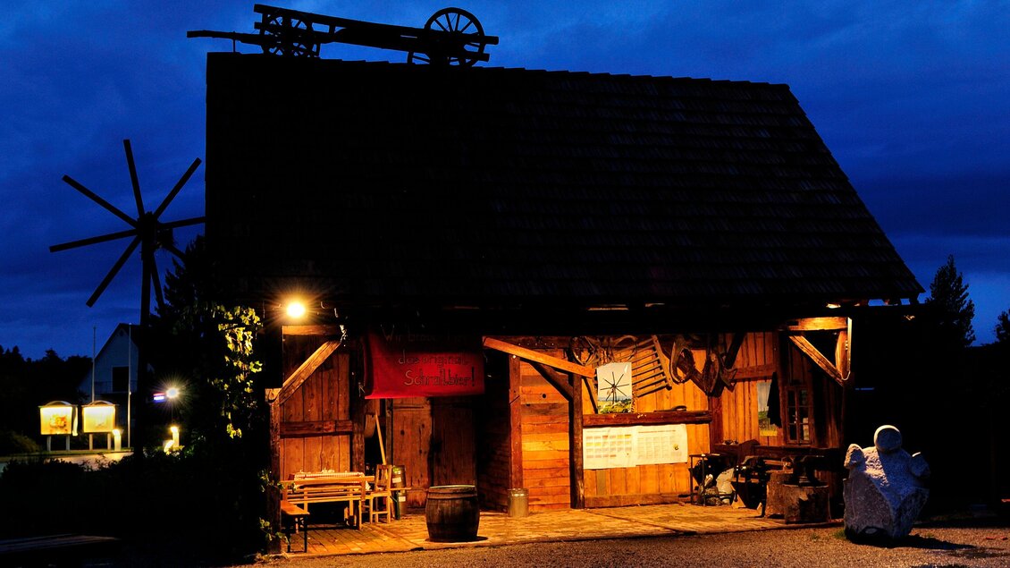 Ein rustikales Holzgebäude bei Nacht, beleuchtet von warmem Licht. Im Hintergrund ist ein Windrad und der Himmel ist dunkelblau. | © SPhotography