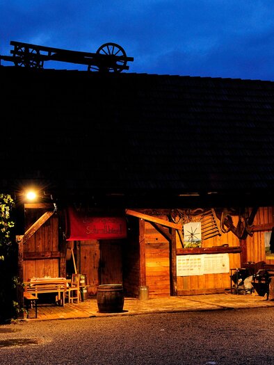 Ein rustikales Holzgebäude bei Nacht, beleuchtet von warmem Licht. Im Hintergrund ist ein Windrad und der Himmel ist dunkelblau. | © SPhotography