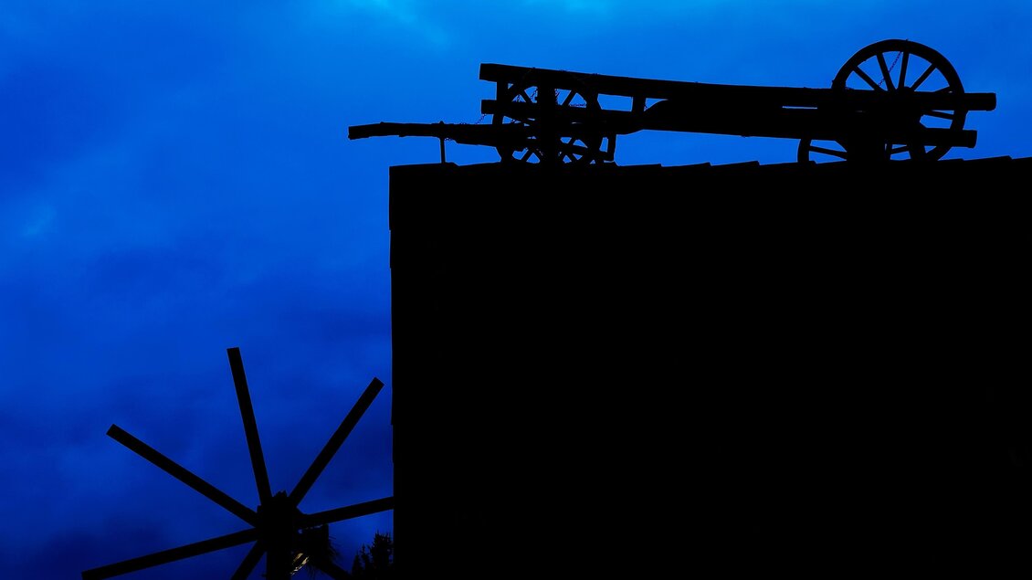 Silhouette eines alten Wagens auf einem Gebäude vor einem moody blauen Himmel. Im Vordergrund ist das Rad eines Windrades zu sehen. | © SPhotography