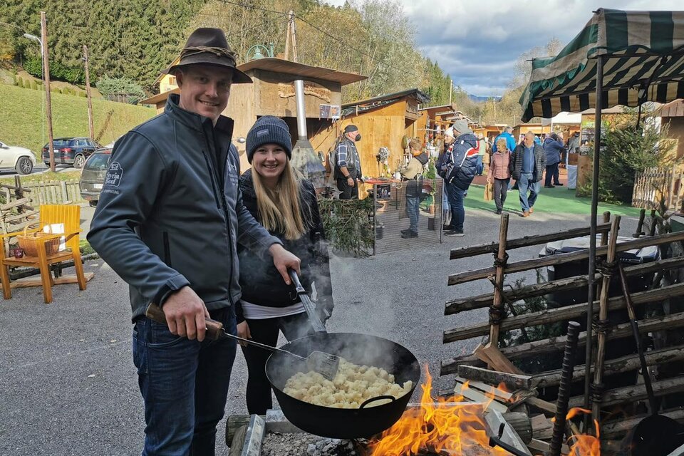 Ein Paar kocht am Feuer in einem Holzpfanne. Im Hintergrund sind Stände und Menschen auf einem Markt zu sehen. | © Stadtgemeinde Frohnleiten