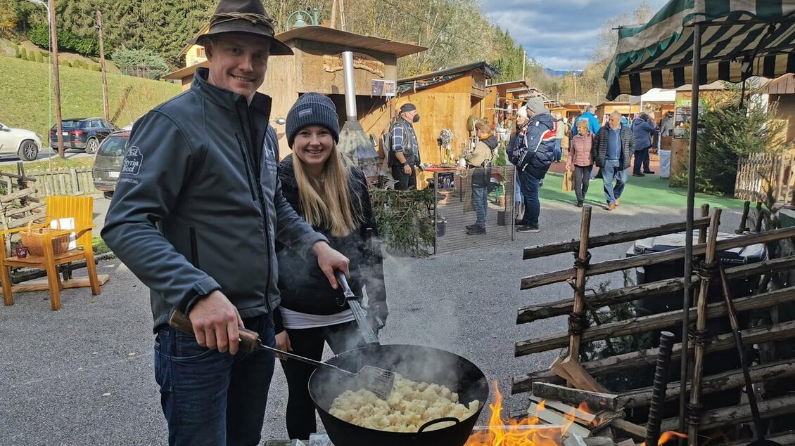 Ein Paar kocht am Feuer in einem Holzpfanne. Im Hintergrund sind Stände und Menschen auf einem Markt zu sehen. | © Stadtgemeinde Frohnleiten