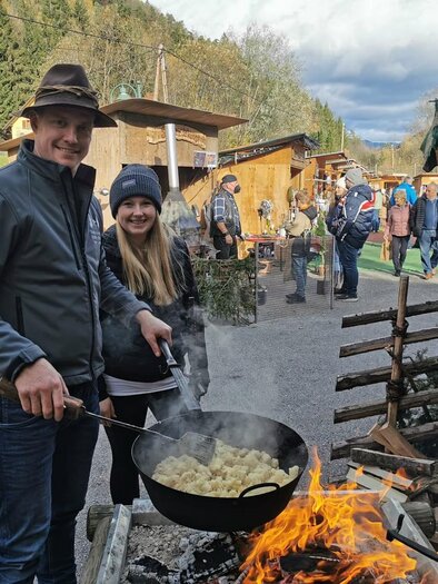 Ein Paar kocht am Feuer in einem Holzpfanne. Im Hintergrund sind Stände und Menschen auf einem Markt zu sehen. | © Stadtgemeinde Frohnleiten