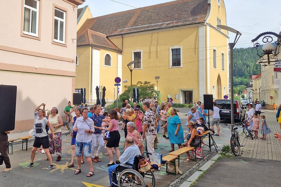 Eine lebhafte Straßenfeier mit vielen Menschen, die tanzen und Spaß haben. Im Hintergrund sind bunte Gebäude und Bäume zu sehen.