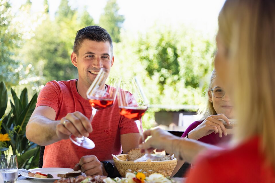 Eine fröhliche Zusammenkunft im Freien mit einer Person, die mit einem Glas anstößt. Im Hintergrund sind frisches Grün und weiterer Genuss zu sehen. | © TVB Südsteiermark – Achromatic Photography