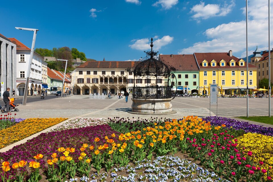 Ein hübscher Platz mit einem Brunnen und bunten Blumenbeeten. Im Hintergrund sind pastellfarbene Gebäude und ein blauer Himmel zu sehen. | © Harry Schiffer
