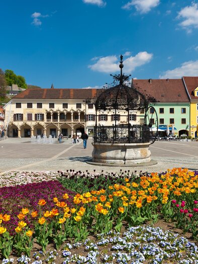 Ein hübscher Platz mit einem Brunnen und bunten Blumenbeeten. Im Hintergrund sind pastellfarbene Gebäude und ein blauer Himmel zu sehen. | © Harry Schiffer