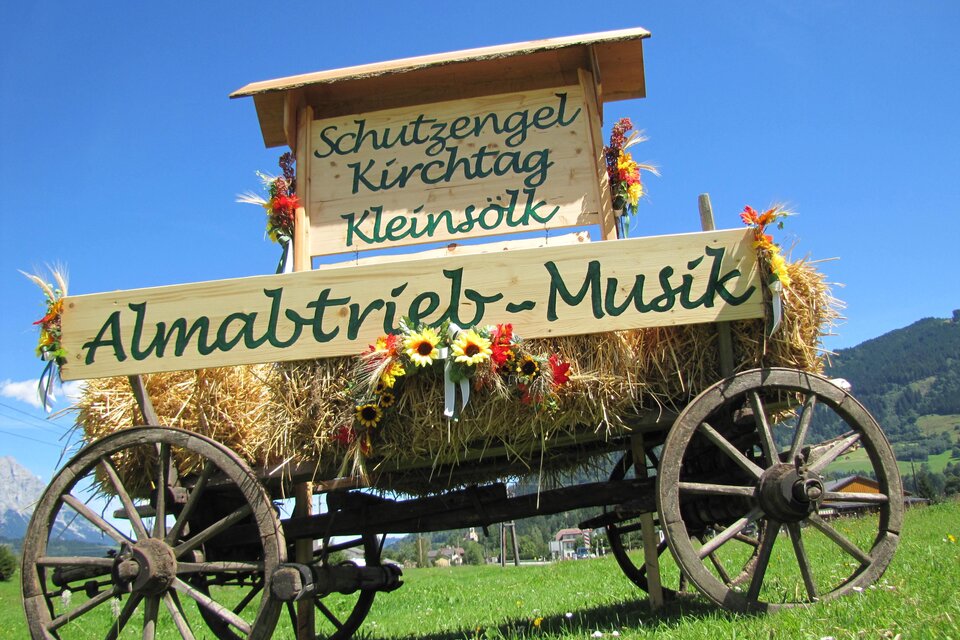 Ein dekorierter Wagen mit Heu und Blumen steht auf einer Wiese. Auf dem Schild steht "Almabtrieb-Musik" und weitere Veranstaltungshinweise.