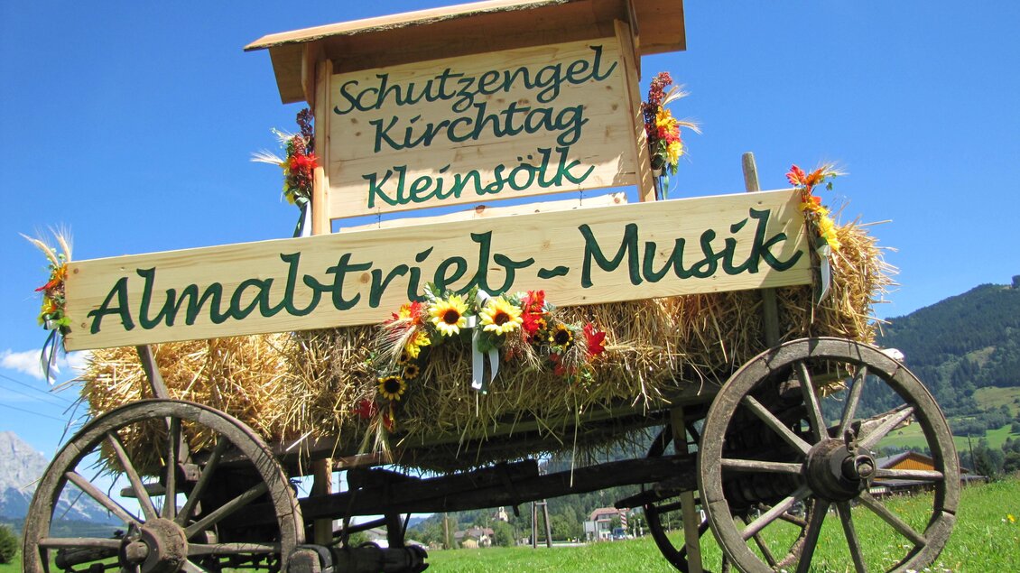 Ein dekorierter Wagen mit Heu und Blumen steht auf einer Wiese. Auf dem Schild steht "Almabtrieb-Musik" und weitere Veranstaltungshinweise.