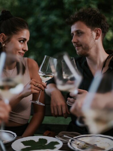 Ein entspanntes Abendessen im Freien mit zwei Personen, die auf Wein anstoßen. Im Hintergrund sind Essensgerichte und eine grüne Umgebung zu sehen. | © Lukas Elsneg