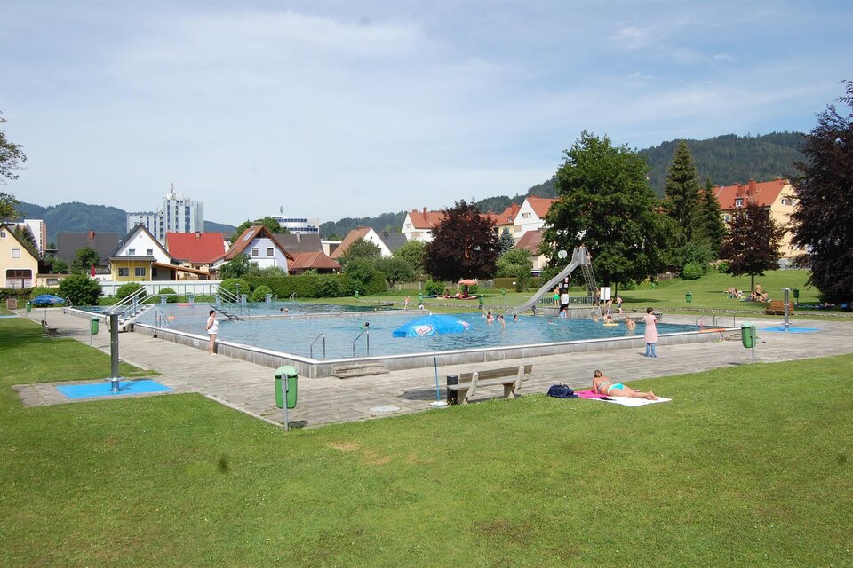 Ein Schwimmbad in einem grünen Park mit ein paar Leuten. Im Hintergrund sind Häuser und Berge sichtbar. | © Freibad Fohnsdorf