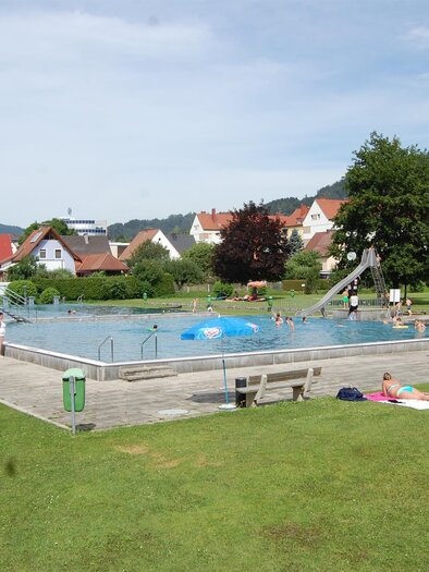 A swimming pool in a green park with a few people. In the background, houses and mountains are visible. | © Freibad Fohnsdorf