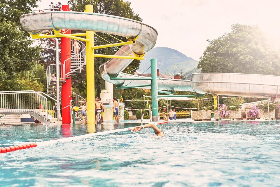 Ein Schwimmbad mit Wasserrutschen und vielen Kindern. Im Hintergrund sind Bäume und Berge zu sehen. | © Sattler Tom