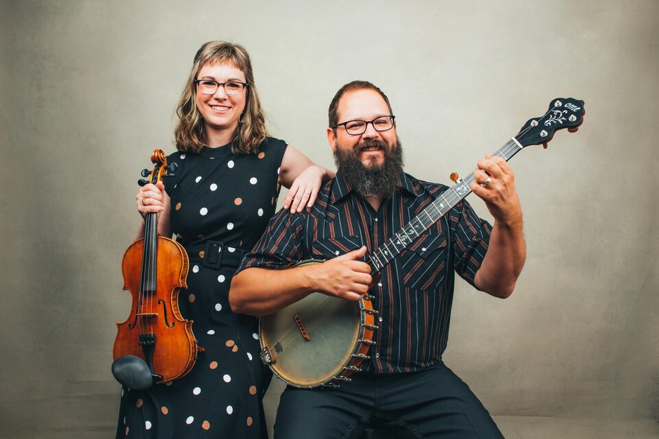 Ein Musikerpaar posiert zusammen vor einem neutralen Hintergrund. Die Frau hält eine Violine, während der Mann eine Banjo spielt und lächelt. | © Sandlin Gaither