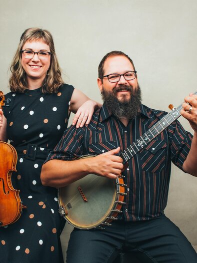 Ein Musikerpaar posiert zusammen vor einem neutralen Hintergrund. Die Frau hält eine Violine, während der Mann eine Banjo spielt und lächelt. | © Sandlin Gaither