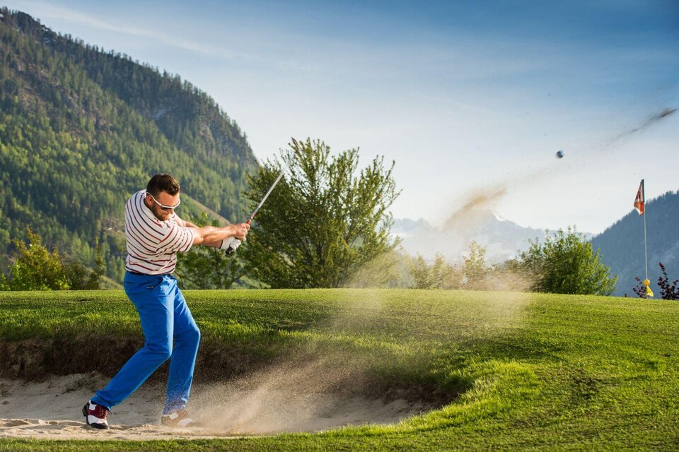 Ein Golfer schlägt einen Ball aus einem Sandbunker beim Golfplatz in Bad Aussee. Im Hintergrund ist die grüne Landschaft des Ausseerlandes mit den Bergen der Umgebung zu sehen. | © MONDI Hotels & Resorts