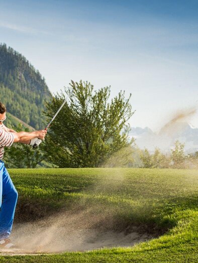 A golfer hits a ball out of a sand bunker at the golf course in Bad Aussee. In the background, the green landscape of the Ausseerland region with the surrounding mountains can be seen. | © MONDI Hotels & Resorts