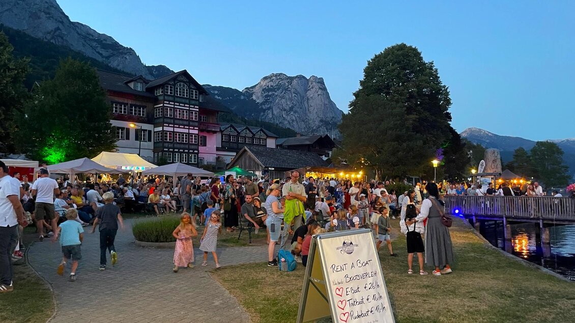 Eine lebhafte Menschenmenge am Abend vor malerischen Gebäuden und Bergen. Stimmungsvolle Lichter und ein See schaffen eine festliche Atmosphäre. | © Bettina Scheck