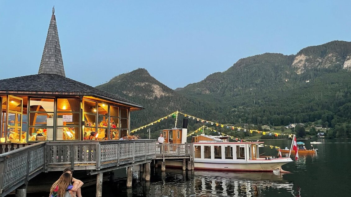 Ein malerischer Holzsteg mit einem Restaurant und einem Boot am Wasser. Im Hintergrund erheben sich grüne Berge unter einem hellen Himmel. | © Bettina Scheck