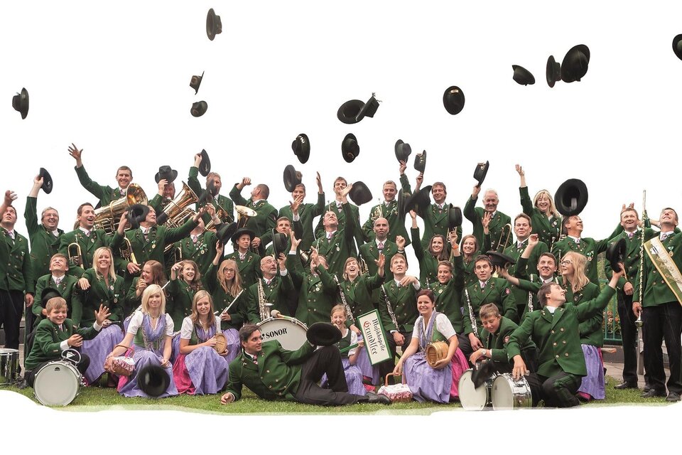 Eine fröhliche Blasmusikgruppe in traditioneller Tracht steht zusammen und wirft ihre Hüte in die Luft. Die Mitglieder sind in grünen Jacken und farbigen Röcken gekleidet und strahlen Freude aus. | © Hannes Grill