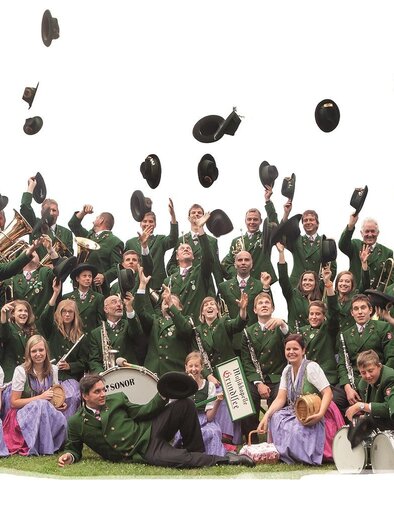 A cheerful brass band in traditional attire stands together and throws their hats into the air. The members are dressed in green jackets and colorful skirts and radiate joy. | © Hannes Grill