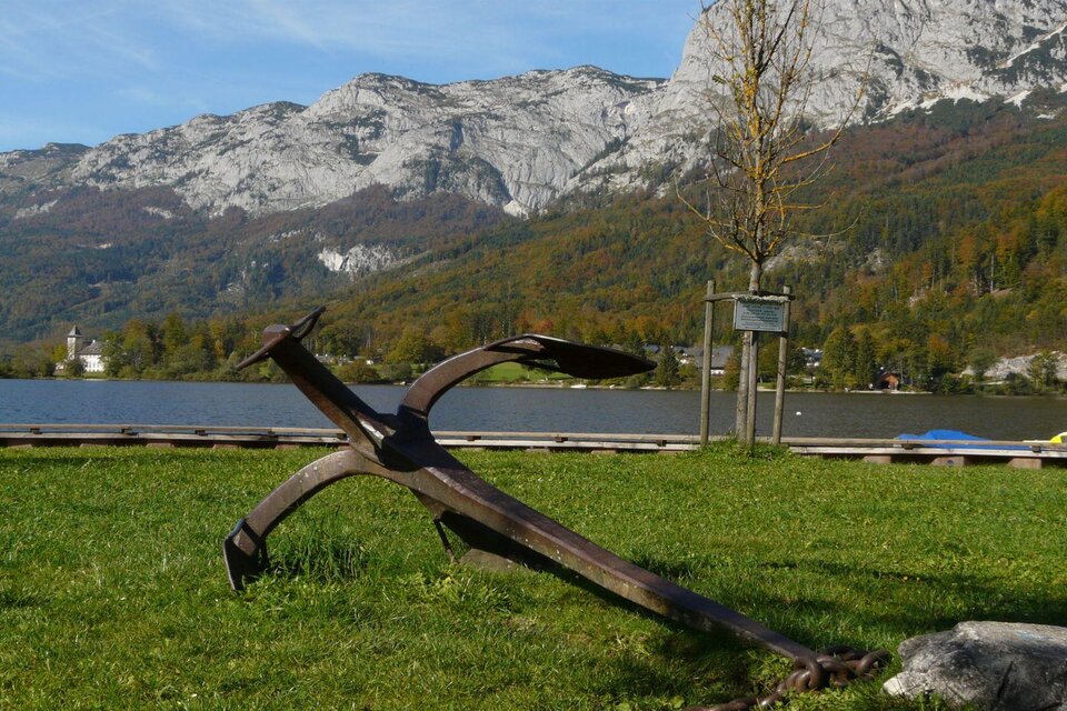 Ein großer Anker liegt auf einer Wiese am Ufer eines ruhigen Sees. Im Hintergrund sind majestätische Berge und Bäume zu sehen. | © TVB Ausseerland - Salzkammergut/Rastl