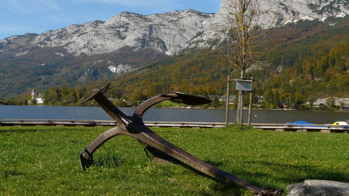 Ein großer Anker liegt auf einer Wiese am Ufer eines ruhigen Sees. Im Hintergrund sind majestätische Berge und Bäume zu sehen. | © TVB Ausseerland - Salzkammergut/Rastl