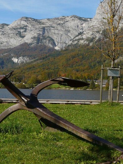 A large anchor lies on a meadow by the shore of a calm lake. In the background, majestic mountains and trees can be seen. | © TVB Ausseerland - Salzkammergut/Rastl