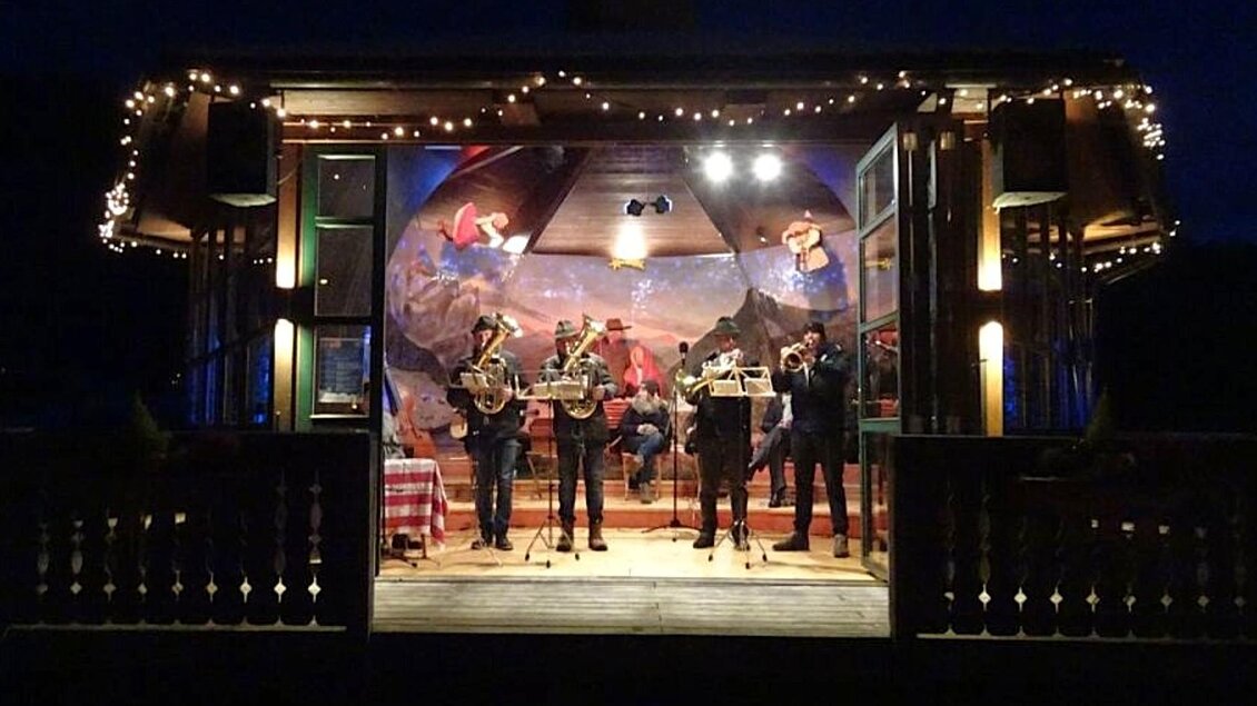 Eine Band spielt auf einer Bühne im Freien, umgeben von bunten Lichtern. Im Hintergrund sind farbenfrohe Wandmalereien zu sehen. | © Mares Rastl