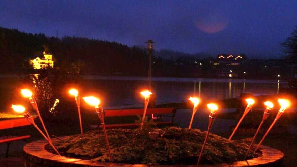 Ein romantischer Abend mit Fackeln, die um einen runden Steintisch angeordnet sind. Im Hintergrund glitzert der See in der Dämmerung. | © Gemeinde Grundlsee
