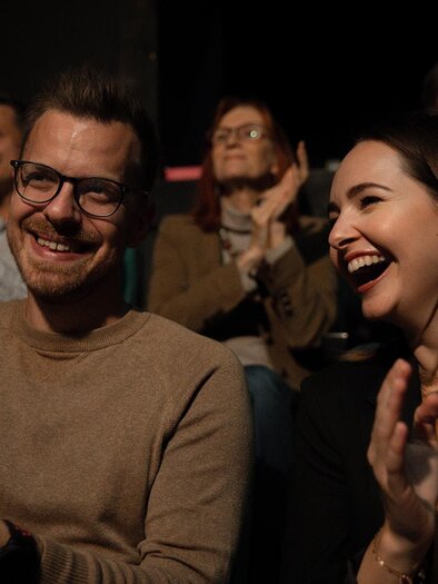 Ein fröhliches Paar sitzt im Publikum und applaudiert. Beide lachen und genießen die Atmosphäre der Veranstaltung. | © achromaticphotography