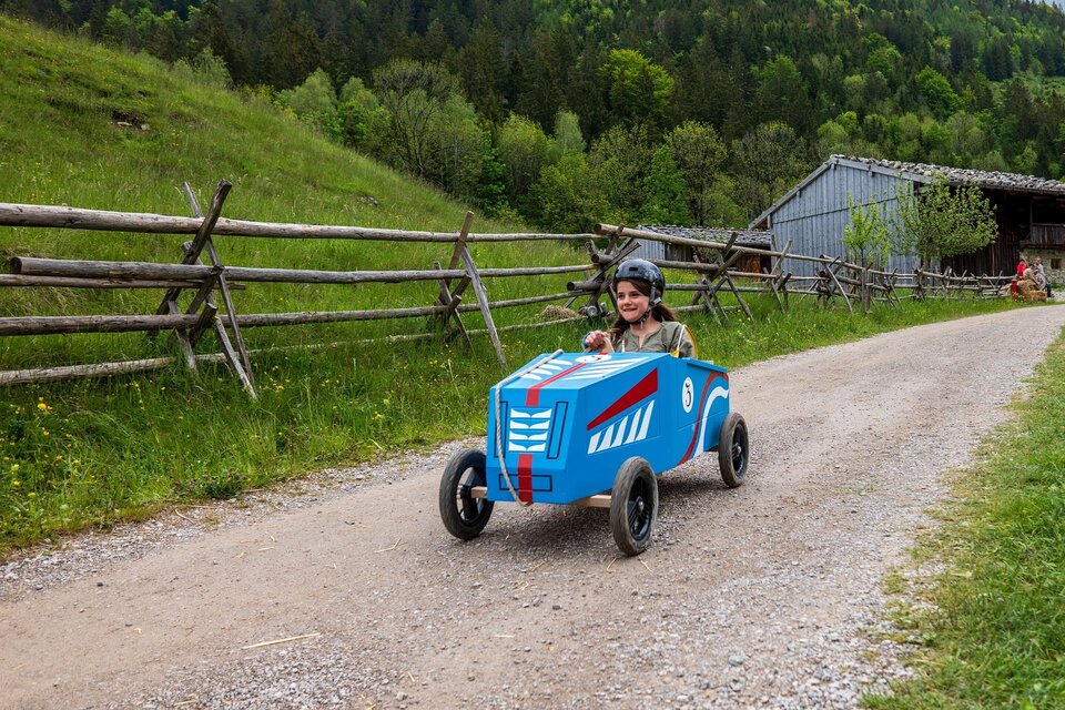 Ein Junge fährt in einem blauen Spielzeugauto auf einem Schotterweg. Im Hintergrund sind grüne Wiesen und eine Scheune zu sehen. | © Pixabay