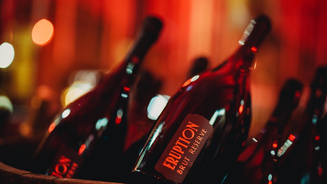 Eine Auswahl an Weinflaschen mit dem Etikett "Eruption Brut Reserve". Die Flaschen sind in einem Körbchen angeordnet und von warmem Licht umgeben. | © Karl Schrotter Photograph