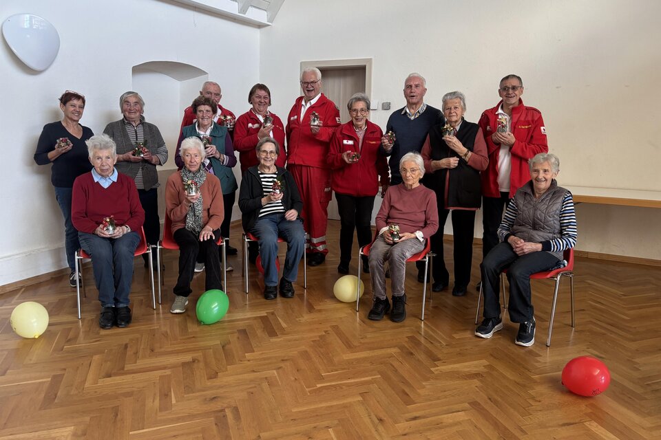 Eine Gruppe von älteren Menschen sitzt und steht in einem hellen Raum. Einige tragen rote Jacken, und es gibt bunte Ballons auf dem Boden. | © Rotes Kreuz Passail
