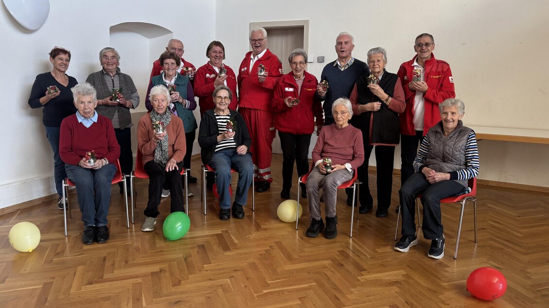 Eine Gruppe von älteren Menschen sitzt und steht in einem hellen Raum. Einige tragen rote Jacken, und es gibt bunte Ballons auf dem Boden. | © Rotes Kreuz Passail