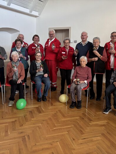 Eine Gruppe von älteren Menschen sitzt und steht in einem hellen Raum. Einige tragen rote Jacken, und es gibt bunte Ballons auf dem Boden. | © Rotes Kreuz Passail