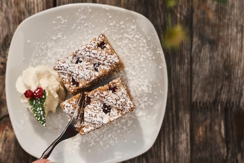 Ein Teller mit zwei Stück Kuchen, bestäubt mit Puderzucker, liegt auf einem Holztisch. Daneben befindet sich eine Portion Sahne und ein Glas mit rohem Getränk. | © TV Gesäuse